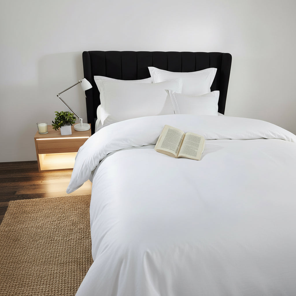 White bedding set on a bed with an open book, lamp, and plant in a room.